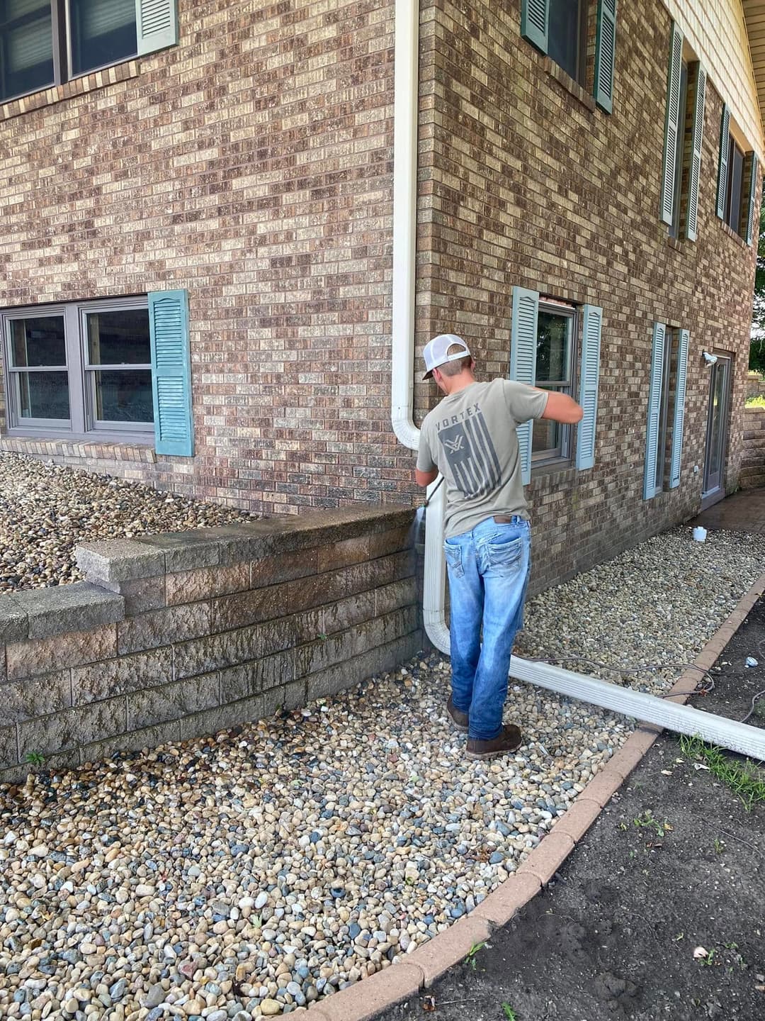 Man in a t-shirt installing a downspout on a brick house exterior with landscaping stones.